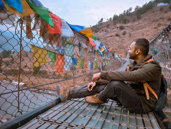 Side view of young man sitting on a suspention bridge with prayer flags outdoors