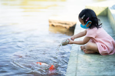 Cute girl wearing protective face mask feeding carp koi fishes at pond.