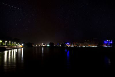 Illuminated buildings by river against sky at night