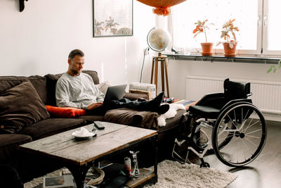 Full length of disabled mature man using laptop while sitting on sofa in living room at home