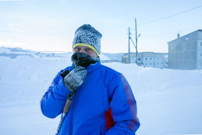 A male skier is standing on a winter street in front of residential buildings on a very frosty day.