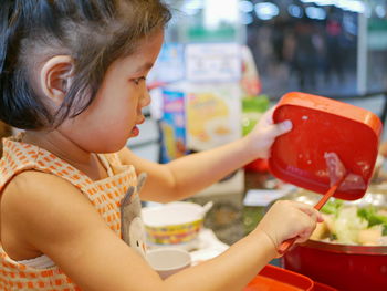 Cute girl holding container and raw chicken with chopstick while preparing food in kitchen