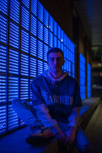 Portrait of serious young man sitting by illuminated building at night