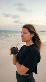 Side view of young woman looking at beach against sky during sunset