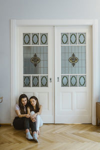 Sad friends sitting on floor together against white door at home