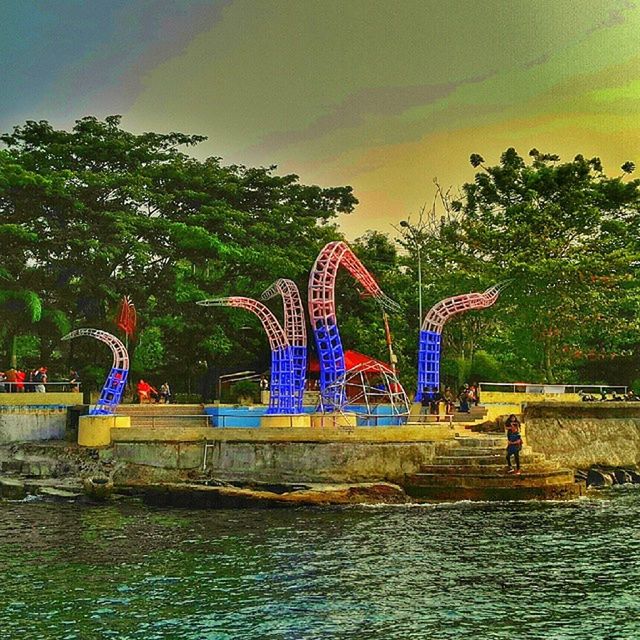 water, art, art and craft, tree, creativity, built structure, sculpture, sky, waterfront, blue, statue, animal representation, human representation, multi colored, park - man made space, architecture, nature, green color, outdoors