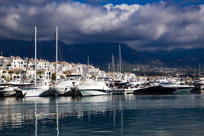 Boats moored at harbor in city against cloudy sky