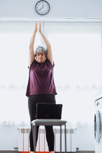 Senior female in activewear watching tutorial video on tablet and raising arms during online fitness training in daytime at home