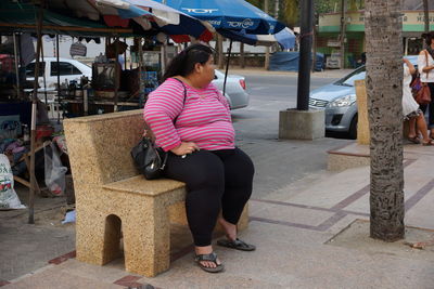 Overweight woman sitting on stone bench at sidewalk in city