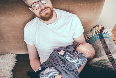 High angle view of man sleeping while holding baby boy on sofa