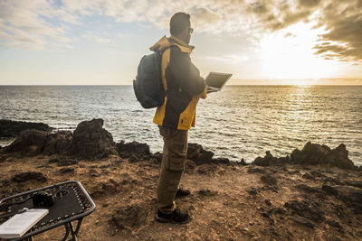 Side view of backpack man using laptop by sea against cloudy sky