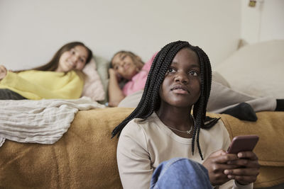 Contemplative teenage girl holding smart phone with female friends lying on bed in background at home