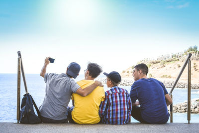 Rear view of friends taking selfie while sitting on staircase at sea