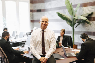 Portrait of smiling working senior male lawyer sitting on desk in office