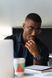 Serious black adult male in formal clothes and eyeglasses sitting at table while taking notes in notebook from papers near netbook in bright workplace