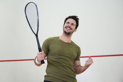 Low angle view of happy mid adult man holding racket while standing by wall in court