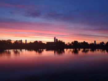 Silhouette buildings by lake against sky during sunset