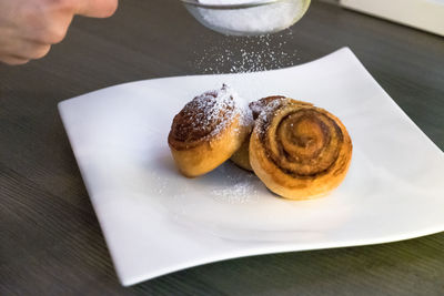 Cropped image of hand sprinkling icing sugar on cinnamon rolls at table