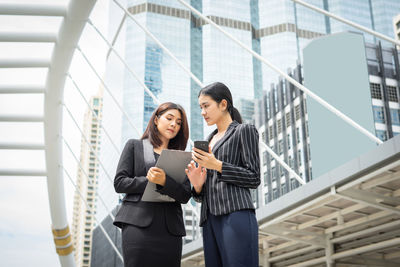 Businesswomen with folder and phone discussing while against buildings in city 