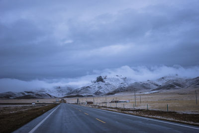 Country road leading towards snowcapped mountains against sky