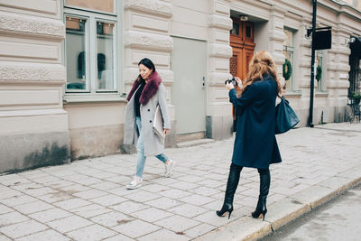 Rear view of woman photographing female friend walking on sidewalk against building in city