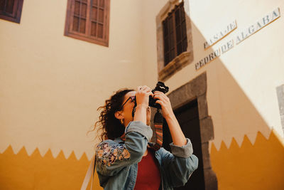 Rear view of young woman photographing while standing against building