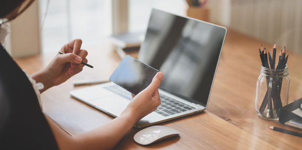 Midsection of woman using mobile phone over laptop on table