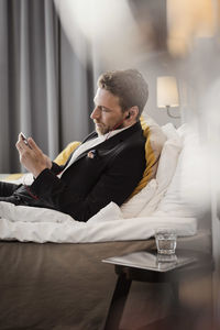 Side view of mature businessman using mobile phone while lying on bed at hotel room