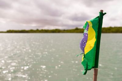 Torn brazilian flag by lake against sky