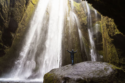 Back view of unrecognizable tourist in warm hooded jacket standing in rocky canyon with powerful picturesque gljufrafoss waterfall with outstretched arms during trip in iceland person