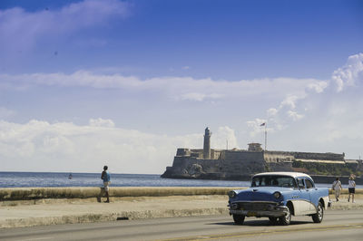 Man standing on car by sea against sky