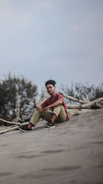 Full length of young man sitting on wood against sky