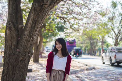 Smiling young woman standing by tree on sidewalk