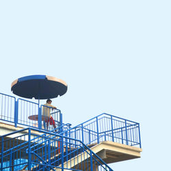 Low angle view of man sitting by railing of staircase against clear sky