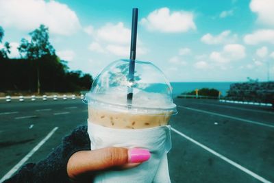 Cropped image of hand holding disposable cup at street against sky