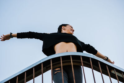 Low angle view of woman with arms outstretched by railing against clear sky