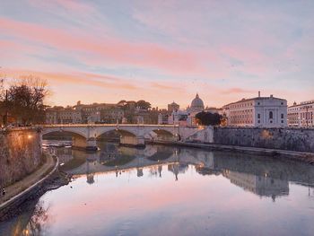 Bridge over river by buildings against sky during sunset