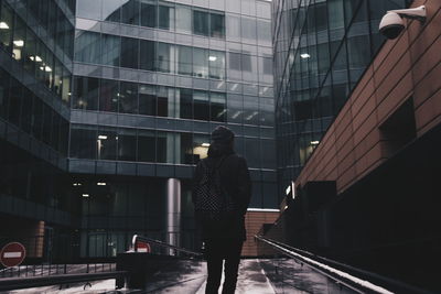 Rear view of person with backpack standing on street against modern buildings during winter