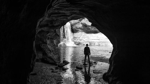 Rear view of man standing at cave entrance against waterfall