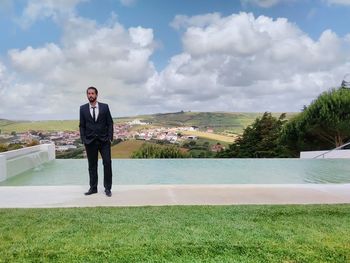 Businessman standing by infinity pool against cloudy sky