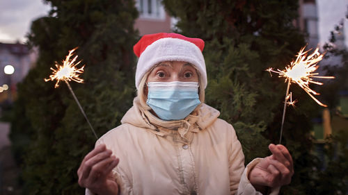 Portrait of senior woman wearing mask holding sparklers against trees