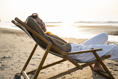 Beautiful young woman relaxing on folding chair at beach during sunset