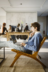 Young businessman using laptop on chair while colleagues in background at creative office