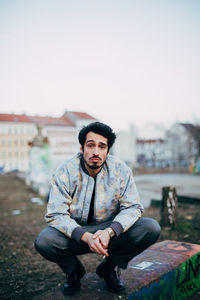 Portrait of young man crouching on retaining wall against sky