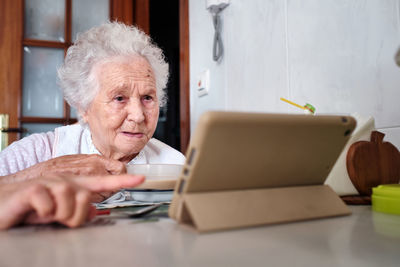 Elderly female with gray hair looking at screen of modern tablet while sitting at table with dish in light kitchen