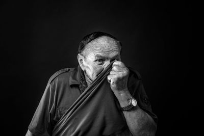 Portrait of senior man hiding face while standing against black background