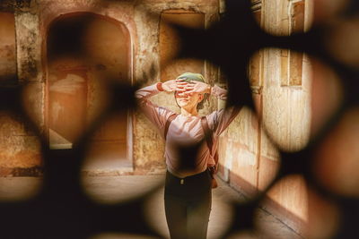 Woman with hands covering eyes standing against wall seen through hole in fence