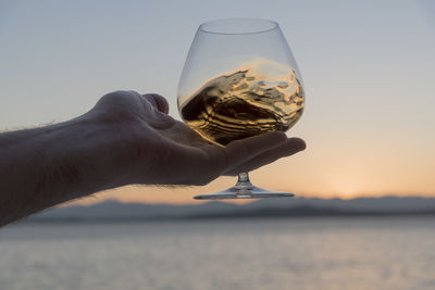 Close-up of hand holding glass against sea during sunset