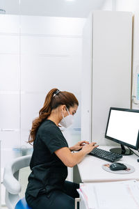 Side view of female doctor in medical mask and uniform sitting at table and working on computer in office