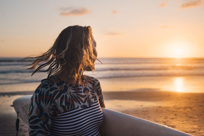 Female surfer with surfboard looking at sea during sunset at beach